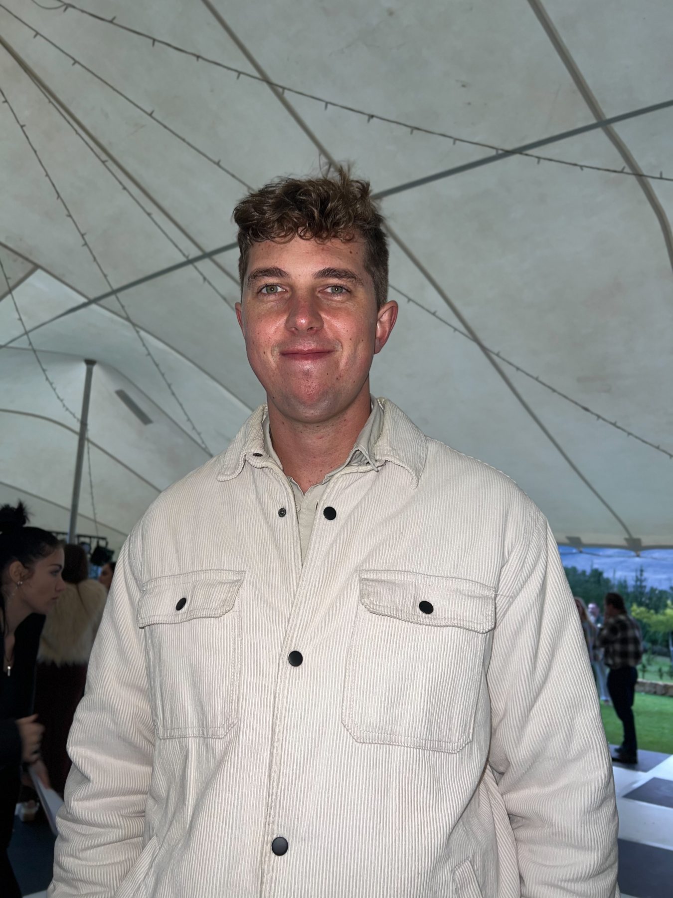 A young man in a cream corduroy jacket under a marquee tent — straight out of camera