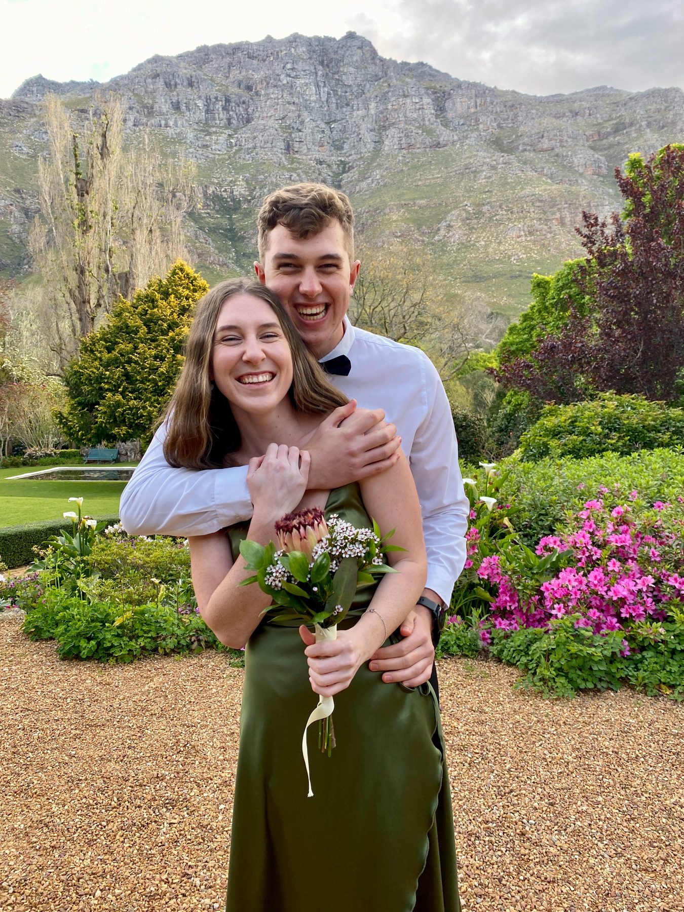 A laughing couple in a mountain garden, she in a green dress holding proteas — straight out of camera